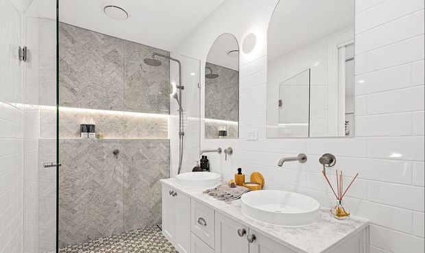 Bright contemporary bathroom with a double vanity, white cabinetry, marble-look benchtop, white subway wall tiles, and a glass-enclosed walk-in shower. The shower features grey veined stone-look wall tiles, a recessed illuminated shelf, and patterned floor tiles. Two vessel basins and brushed metal tapware complete the refined fit-out.