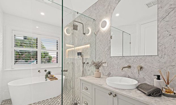 Bright, refined bathroom featuring a freestanding white bathtub, frameless glass shower enclosure, marble-look wall and vanity surfaces, patterned floor tiles, and a pale built-in vanity with vessel basin. White plantation shutters and mirrored cabinetry add a clean, classic finish.
