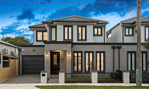 Street-facing view of a contemporary multi-storey townhouse with a low-pitched tiled roof, grey brick lower facade, light rendered upper walls, black-framed windows, an integrated single garage, and a small fenced front garden. Overhead utility wires and neighbouring homes are visible.