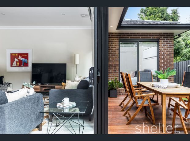A split interior-exterior view showing a contemporary living room opening directly onto a timber-decked outdoor dining area. The living room has pale walls, grey upholstered sofas, a glass coffee table, timber media console and television, while the adjacent patio features a timber dining table and folding chairs, brick walls, dark-framed glazing, and a large roof skylight or opening.