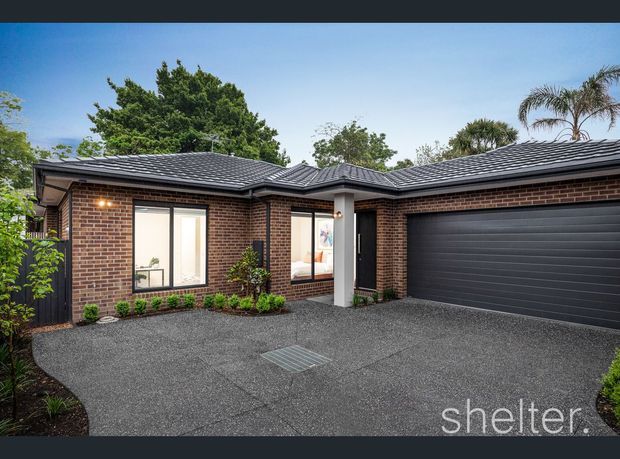 Single-storey residential house facade viewed from the driveway, featuring dark brown face-brick walls, a low-pitched dark tiled roof with multiple hips, black-framed windows and entry door, and a wide dark sectional garage door. The front area is finished with dark textured paving and modest planted garden beds.