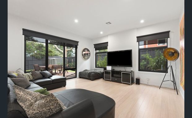 A bright contemporary living room with pale timber flooring, white walls, recessed ceiling lights, large black-framed windows and sliding glass doors, and dark sectional seating. The room includes a media unit with television, a freestanding tripod floor lamp, and views to a small outdoor deck and garden beyond the glazing.