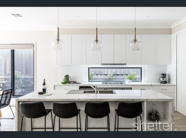 Bright contemporary kitchen with a large white island and integrated seating for four, white cabinetry, pale stone benchtops, stainless-steel appliances, and three clear glass pendant lights. A dining area and glazed doors are visible at the left, with a small courtyard or exterior view beyond the rear window.