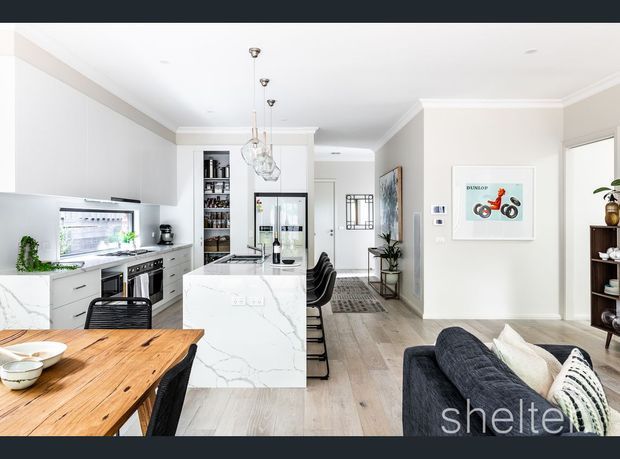 Bright open-plan kitchen, dining and living area with white shaker-style cabinetry, a large white marble-look island, timber dining table, pale timber flooring, pendant lights and a dark sofa. The kitchen includes integrated appliances, open shelving and a glass-fronted drinks cabinet, while the adjoining living area and hallway are visible beyond.