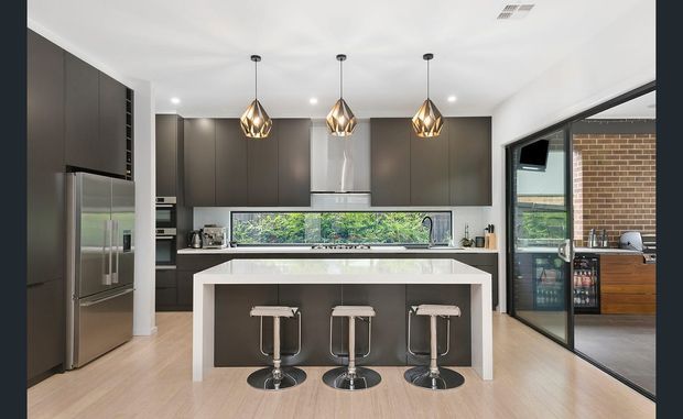 Contemporary open-plan kitchen with dark matte cabinetry, a large white island with three stools, stainless-steel appliances, timber-look flooring, pendant lights, and a glazed opening to an adjacent outdoor or living area.