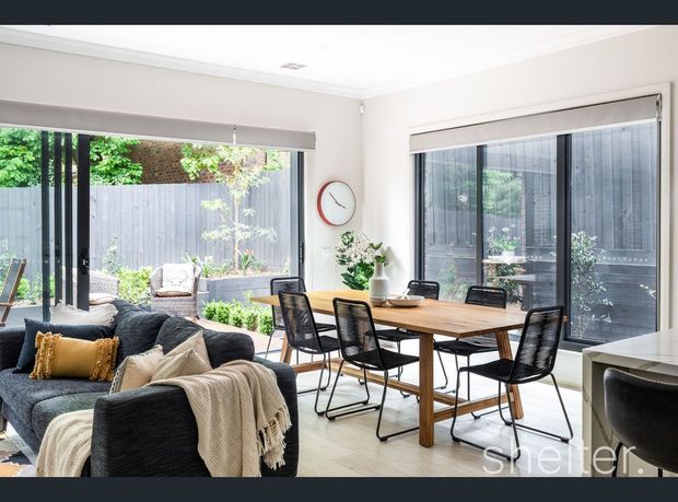 Bright open-plan living and dining area with large glazed doors and windows opening to a small planted courtyard. A dark upholstered sofa with cushions and throw occupies the foreground, while a timber dining table with black chairs sits beside the glazing. Neutral walls, light flooring, roller blinds, and simple contemporary furnishings create an airy, inviting interior.