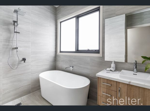 Modern bathroom featuring large-format grey wall and floor tiles, a freestanding white bathtub, wall-mounted shower fittings, a white vanity basin with timber-look cabinetry, a large mirror, and a black-framed window. The room is bright and clean with contemporary fixtures and finishes.