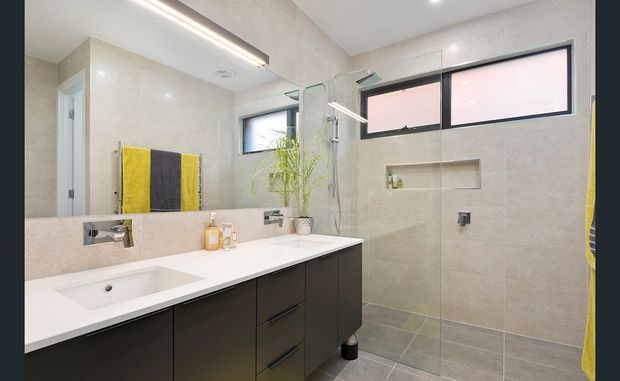 Contemporary bathroom with a long white vanity and integrated basin, dark built-in cabinetry, large-format beige wall and floor tiles, a frameless glass walk-in shower, recessed shelving, and high horizontal windows. A wall mirror reflects the vanity and bright linear ceiling lighting.