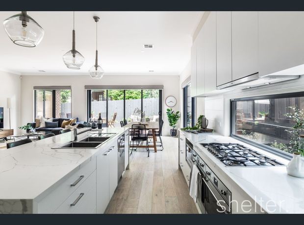 Bright contemporary open-plan kitchen with a large white stone island, extensive white cabinetry, integrated appliances, gas cooktop, and pendant lighting. A dining area and living room are visible beyond the kitchen, with large windows bringing in abundant natural light.