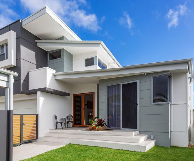 Contemporary multi-level residential facade with layered flat rooflines, projecting upper volumes, crisp white and charcoal finishes, pale horizontal cladding, and a sheltered entry porch. The front elevation includes steps, glazed sliding doors, windows, a small planted pot, lawn, and a slatted side gate.