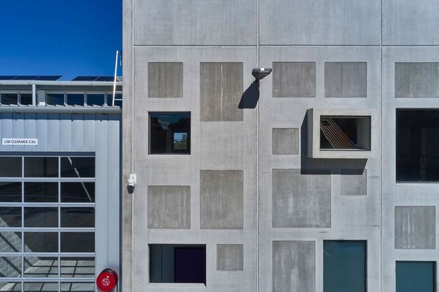 Exterior view of a contemporary institutional or commercial building facade. The facade is composed of large pale grey concrete panels with varied inset rectangular textured panels, small windows, a projecting window box, and dark recessed openings. An adjacent glazed industrial-style structure with a low-clearance sign is visible at the left edge.
