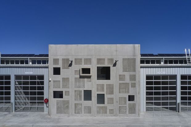 Front-facing exterior view of a contemporary industrial or commercial building. A large patterned concrete central facade panel with varied inset rectangular openings and panels is flanked by metal-clad wings with tall glazed sectional doors. Solar panels are visible along the roofline beneath a clear blue sky.
