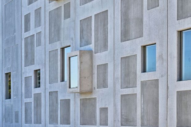Close-up exterior view of a contemporary concrete facade composed of pale grey precast or cast concrete panels with a geometric arrangement of recessed and projecting rectangular elements. Narrow rectangular windows are integrated into the facade, including a projecting box-like window surround. The image is architectural and materially minimal, with no visible interior or surrounding landscape.