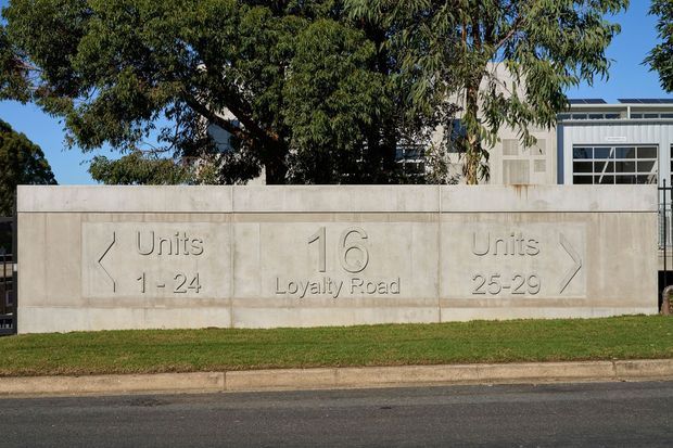 Street-facing architectural entry sign for a multi-unit residential development. A broad pale concrete wall has engraved directional arrows and lettering reading “Units 1–24”, “16 Loyalty Road”, and “Units 25–29”. Mature trees, lawn, curb, and asphalt street frame the sign, with part of a contemporary building visible behind it.
