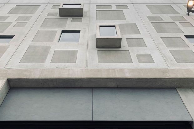 Close-up upward-looking view of a minimalist concrete facade featuring a regular grid of recessed rectangular panels and projecting box-like window surrounds. The pale grey architectural surface has crisp geometric detailing, with dark glazing visible in some openings and a linear dark element along the lower edge.