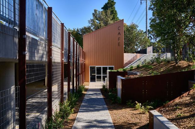 Exterior view of a contemporary café building and landscaped access path. The café is clad in rust-coloured corrugated metal, with vertical signage, glazed entry doors, and a contrasting tall metal-mesh facade or enclosure on the left. Steel retaining walls, concrete steps, gravel, and planted garden beds create a modern institutional or commercial setting beneath mature trees and visible overhead utility lines.