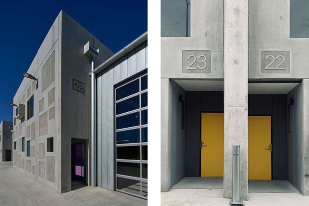 A split view of a contemporary industrial-style building facade. The left side shows a tall raw concrete and corrugated metal elevation with a large glazed sectional garage door, small inset windows, a dark pedestrian entry, and the number 28. The right side shows two recessed entrances with vivid yellow doors, exposed concrete surrounds, small upper windows, and address numbers 23 and 22. The composition emphasizes commercial or service-building frontage and robust geometric materials.