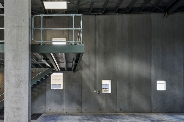 Industrial interior with tall raw concrete walls, exposed structural ceiling services, and a metal stair leading to a small mezzanine platform with utilitarian guardrails. Small square windows punctuate the wall, while the concrete floor and dark steel elements create a stark, functional atmosphere.