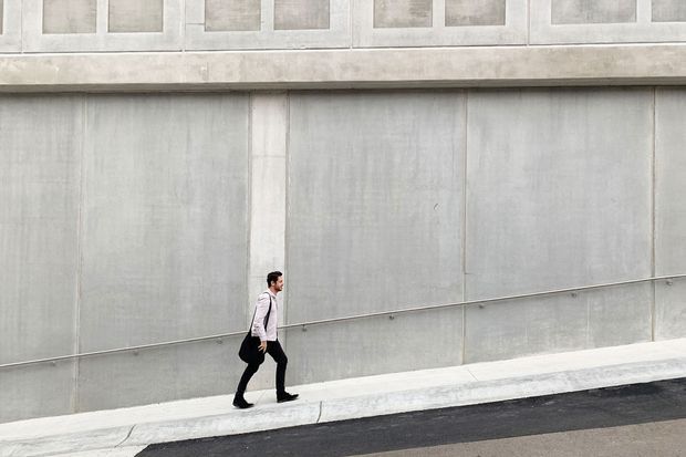 Minimalist urban architectural exterior showing a tall, pale grey concrete or cement-panel facade with large vertical panel divisions, a projecting horizontal ledge, and a row of small square recessed openings near the roofline. A narrow concrete sidewalk and asphalt street run along the base, with a pedestrian providing scale.