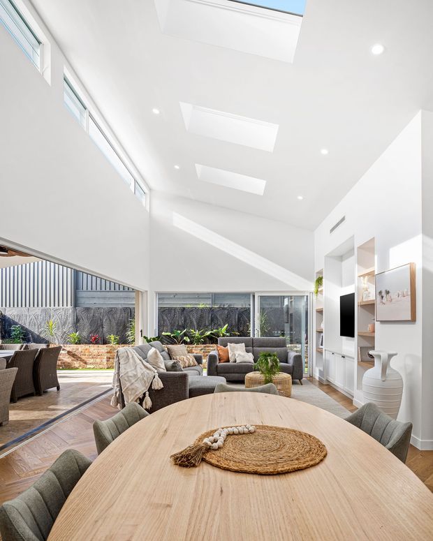 Bright open-plan living and dining space with a large light-wood dining table in the foreground, upholstered dining chairs, a neutral sectional sofa, and built-in white shelving with television. The room features extensive glazing, a very high white ceiling with multiple skylights, recessed lighting, and views to a landscaped courtyard with a brick garden wall. Natural woven and timber accents create a relaxed contemporary atmosphere.