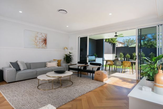 Bright contemporary open-plan living room opening through large sliding glass doors to a covered outdoor dining area and landscaped courtyard. The room features a grey sectional sofa, round nesting coffee tables, textured area rug, white walls with decorative moulding, recessed lighting, and warm herringbone timber flooring. Indoor plants and neutral furnishings create a refined, inviting atmosphere.