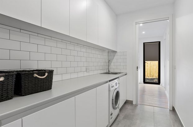 A bright, narrow contemporary laundry room with white full-height cabinetry, a long light-grey benchtop, white subway-tile splashback, and integrated front-loading washing machine. Woven black storage baskets sit on the counter. A doorway leads to a timber-floored exterior or adjoining area.