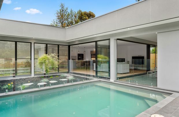 Modern single-storey home arranged around a private courtyard pool. The image shows a crisp white flat-roofed exterior with extensive black-framed floor-to-ceiling glazing, a turquoise swimming pool, gravel landscaping, and views into an open-plan kitchen and living area.