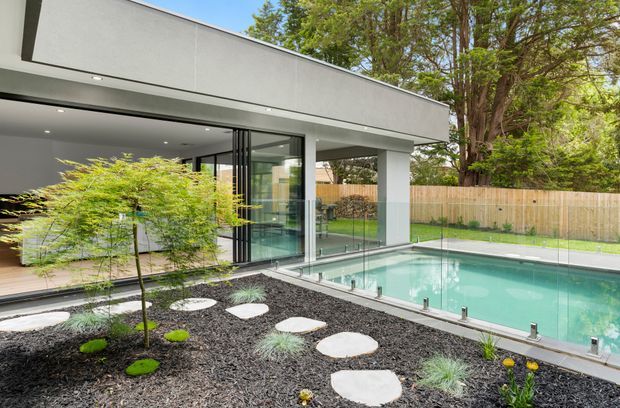 Contemporary flat-roofed house with expansive floor-to-ceiling glazing opening onto a swimming pool. The image shows a minimalist landscaped courtyard with dark gravel, stepping stones, ornamental planting, and a timber privacy fence, surrounded by mature trees.