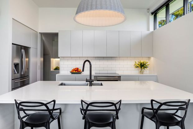 Bright contemporary kitchen with a large white island, integrated sink and dark gooseneck tap, three black bar stools, white subway-tile splashback, handleless white cabinetry, stainless-steel appliances, and high clerestory-style windows. A large grey pendant light hangs above the island.