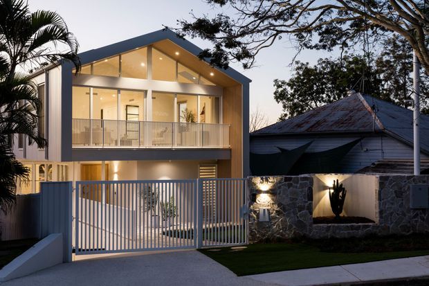 Twilight exterior view of a contemporary multi-storey residence with a steeply pitched asymmetrical roof, extensive upper-level glazing, warm interior lighting, and a front balcony. A modern rendered and timber-toned facade contrasts with an older corrugated-roof structure and dark stone boundary wall, with a gated driveway in the foreground.