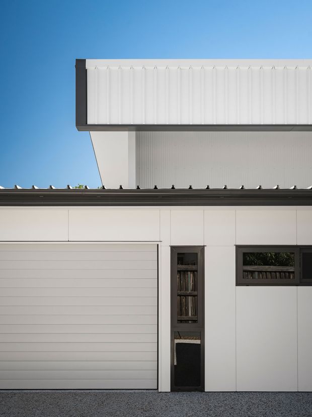 Cropped architectural exterior of a crisp white multi-level residential facade, featuring a sectional garage door, narrow dark-framed glazed entry, horizontal clerestory-style window, dark roof edges, and a projecting upper volume clad in white vertically ribbed panels. The composition is minimalist and geometric against a clear blue sky.