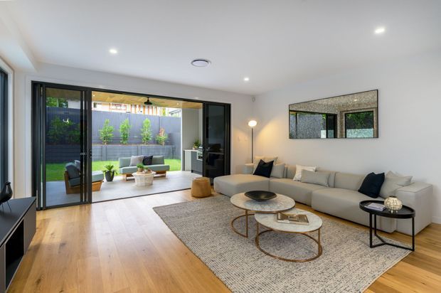 Bright contemporary living room with a large cream sectional sofa, layered round coffee tables, textured area rug, black side tables, and a full-width black-framed glazed opening leading to a landscaped courtyard. Light timber flooring, white walls and recessed ceiling lights create an airy, refined interior.