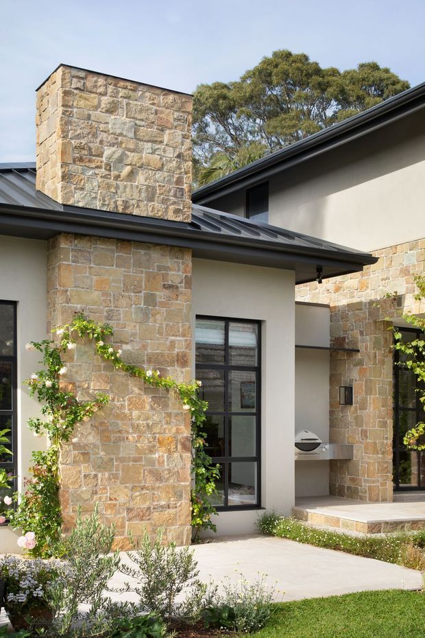 Exterior view of a contemporary single-storey home featuring substantial warm-toned natural stone masonry columns and chimney, smooth light rendered walls, dark metal pitched roof sections, and black-framed glazing. A paved walkway leads through a landscaped garden with climbing plants and low planting. The composition highlights the facade, roofline, chimney, covered entry, and outdoor barbecue area.