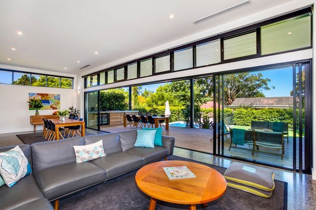 Bright, open-plan contemporary living room with a large grey sectional sofa and round timber coffee table, opening through extensive black-framed glazing onto a timber deck and swimming pool. The space also includes a dining area and outdoor entertaining area visible beyond the glass, with abundant natural light and greenery outside.