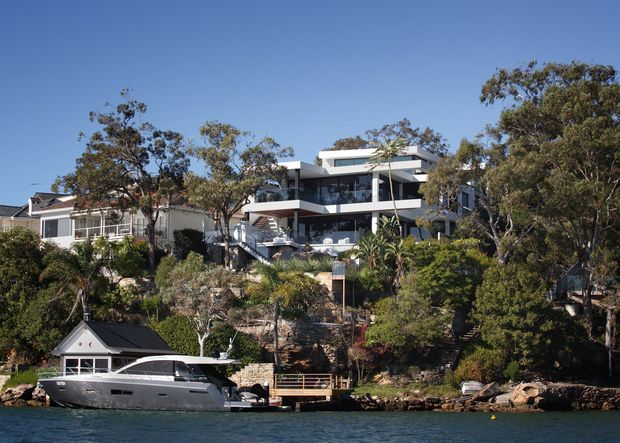 A waterfront view of a contemporary multi-level residence built into a densely landscaped, sloping site. The home features broad horizontal forms, extensive glazing, white and dark facade elements, balconies, and terraces overlooking the water. A neighboring house, shoreline structures, and a moored boat are visible in the foreground.