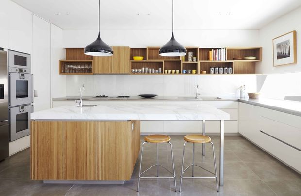 Bright contemporary kitchen with a large central island, white stone surfaces, warm timber cabinetry, open shelving, integrated stainless-steel appliances, and two black pendant lights. The space has a clean, spacious layout with stools at the island and grey tiled flooring.