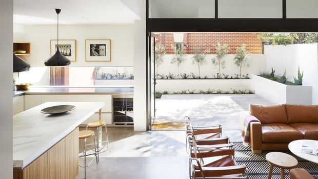 Bright contemporary open-plan kitchen and living space opening onto a landscaped courtyard. The kitchen features a large light stone island with timber cabinetry, integrated appliances and pendant lighting, while the living area has a brown leather sofa, lounge chairs and round coffee tables. Full-height black-framed glazing connects the interior to a paved courtyard with built-in white planters, layered planting and a brick boundary wall.