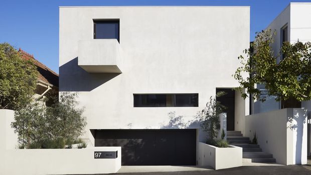 Street-facing view of a contemporary, minimalist two-storey residence with a smooth white rendered facade, strong cubic geometry, recessed dark-framed windows, a projecting upper-level box, and an integrated dark garage. A stepped entry path rises beside low white rendered planter walls, with mature planting softening the facade. A neighbouring tiled-roof house is partially visible at left.