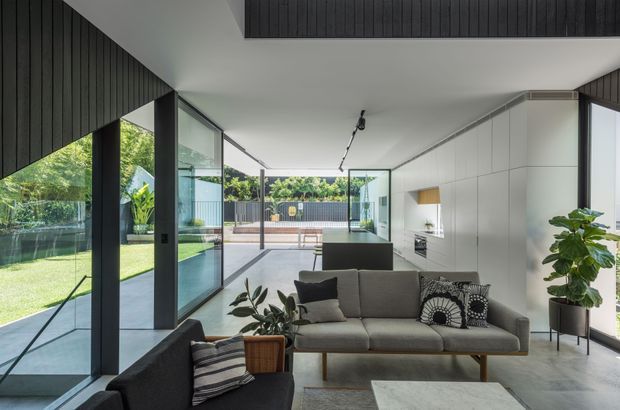 A bright, open-plan contemporary living room with floor-to-ceiling glazing opening onto a landscaped yard and terrace. The room features a grey sectional sofa, dark upholstered armchair, low coffee table, minimalist track lighting, extensive white built-in cabinetry, a dark kitchen island in the background, and dark vertical timber wall and ceiling panelling. The space has a clean, refined indoor-outdoor connection with abundant natural light.
