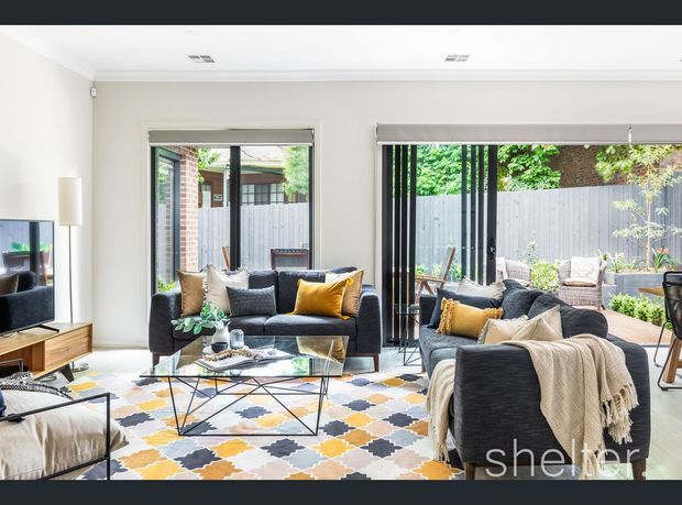 Bright contemporary living room with large glazed openings and black-framed sliding or folding doors opening to a fenced courtyard. The room contains dark sofas with neutral and mustard cushions, a glass coffee table, patterned geometric rug, timber media console, and abundant natural light. A small outdoor seating area is visible beyond the glazing.