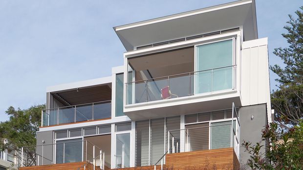 Modern multi-level residential facade with clean white and grey rendered surfaces, extensive glazing, glass balcony balustrades, projecting flat roof forms, external louvres, and warm timber privacy screens or decking. The image is photographed from outside and focuses on the building exterior.