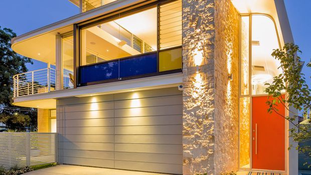 Exterior dusk view of a modern multi-level residence with broad floor-to-ceiling glazing, curved projecting balconies, horizontal metal cladding, a textured stone feature wall, slim metal balustrades, and a vivid orange entry door. Warm interior and facade lighting highlight the contemporary facade.