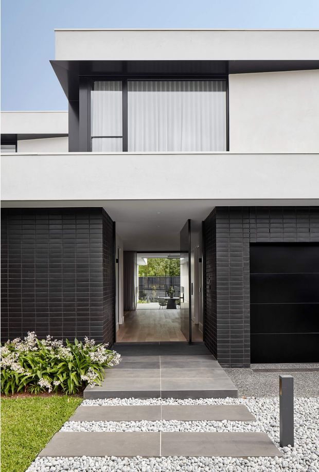 Front exterior of a contemporary two-storey residence with crisp white rendered walls, bold black-framed glazing, dark brick-tiled facade sections, and a black garage door. A recessed entry passage opens through the house toward a landscaped rear courtyard. The approach uses large grey paving slabs separated by white gravel, with minimalist planting and a simple bollard light.