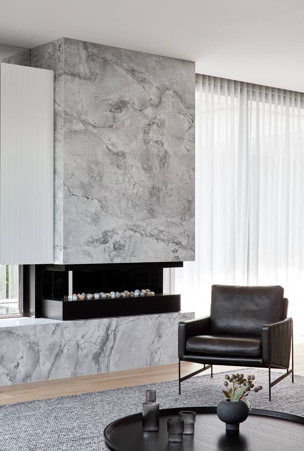 A refined contemporary living room centred on a sculptural marble-clad fireplace and hearth. The room features pale timber flooring, a dark leather lounge chair, a large grey textured rug, sheer full-height curtains, black built-in fireplace detailing, and a round black coffee table with minimal decorative objects. The palette is predominantly cool grey, white, black, and dark brown, with abundant natural light.