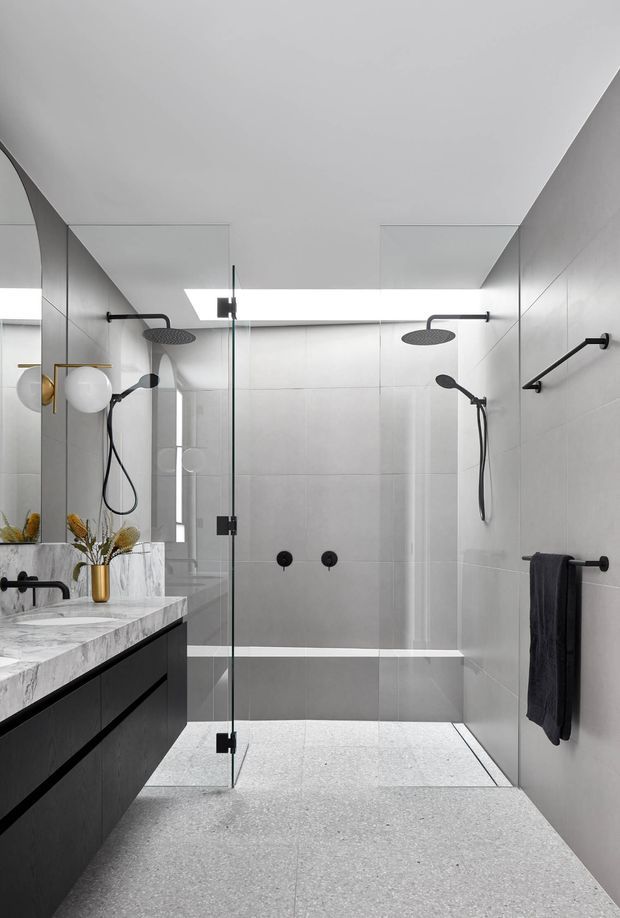 Contemporary bathroom featuring a generous frameless glass walk-in shower with dual rain showerheads and handheld fixtures, pale large-format wall tiles, a terrazzo-like floor, dark floating vanity cabinetry, and a white marble benchtop. Black tapware, towel rails, and shower fittings provide contrast, while a large mirror and sculptural lighting add brightness.
