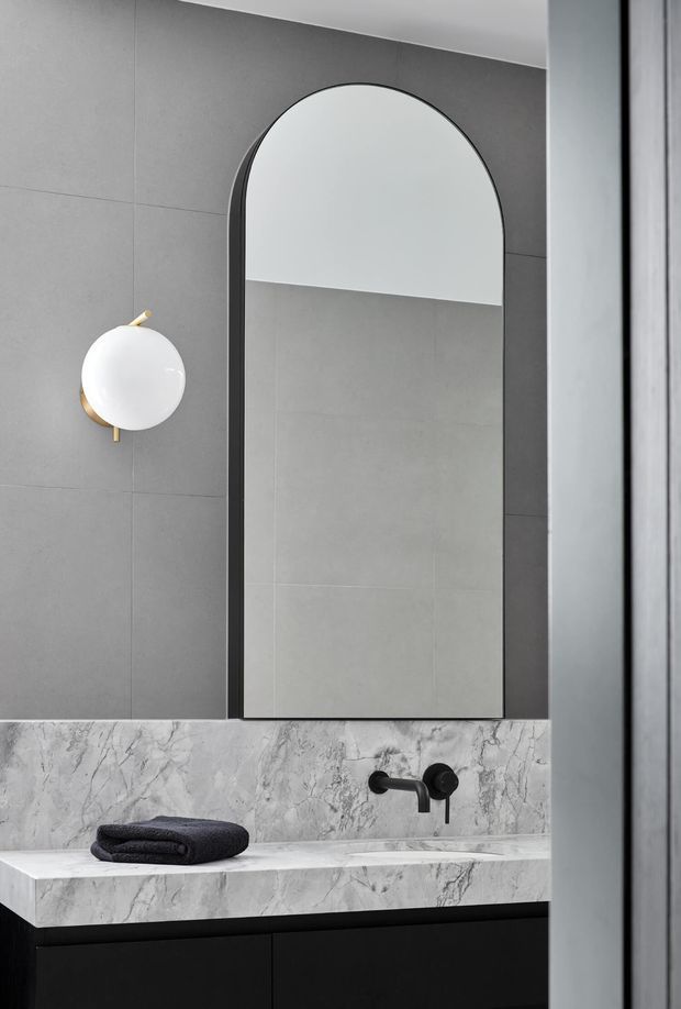 A refined contemporary bathroom vanity featuring a large arched black-framed mirror, grey tiled walls, a veined marble vanity splashback and benchtop, matte black wall-mounted tapware, dark cabinetry, and a spherical white-and-brass wall light. The palette is monochromatic grey, white, and black with subtle warm metallic detailing.