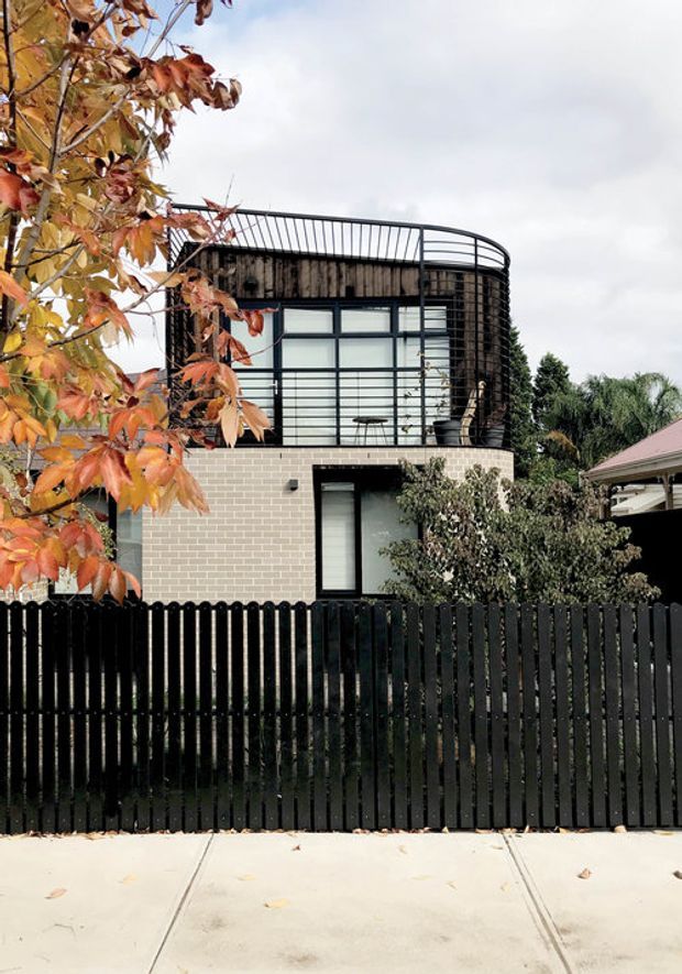 Street-facing view of a contemporary multi-storey urban residence with a pale brick lower level, dark timber or metal-clad upper volume, extensive black-framed glazing, and a curved rooftop or terrace balustrade. A tall black picket fence, mature planting, autumn foliage, and neighbouring homes frame the facade.