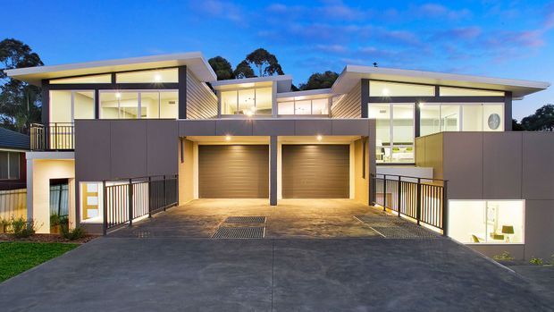 A contemporary multi-level residential development viewed from the street at dusk, featuring two attached homes with flat, projecting roof planes, extensive glazing, dark vertical cladding, pale rendered walls, balconies, and two integrated double garages. A broad concrete driveway and illuminated interiors emphasize the clean modern facade.