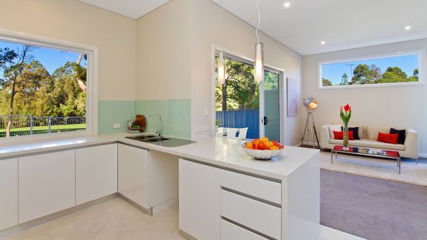 Bright open-plan kitchen and living room with minimalist white cabinetry, a white island bench, pale aqua glass splashback, and light flooring. The adjoining living area has a cream sofa with red and black cushions, a glass coffee table, and large windows overlooking greenery. Recessed ceiling lights and pendant lighting create an airy contemporary interior.