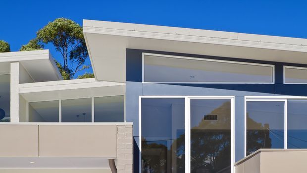 Contemporary residential facade viewed from outside, featuring clean white rooflines and framing, dark blue-grey wall cladding, extensive floor-to-ceiling glazing, and a pale rendered balcony or privacy wall. The image shows a crisp, minimalist composition under a clear blue sky, with trees reflected in the windows.